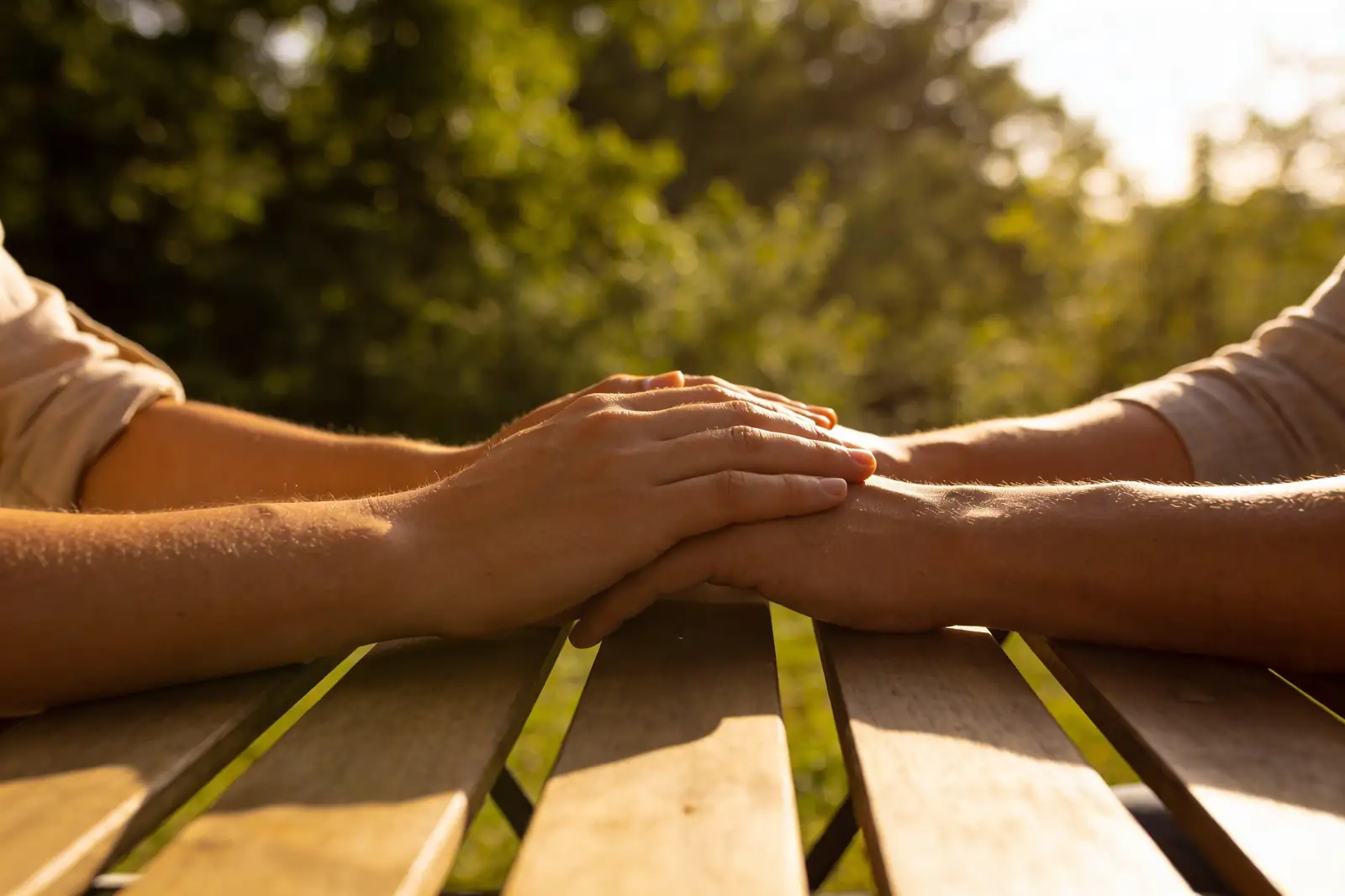 A person sitting with a grieving friend, hands gently held together on a sunlit table — a quiet gesture of comfort and presence