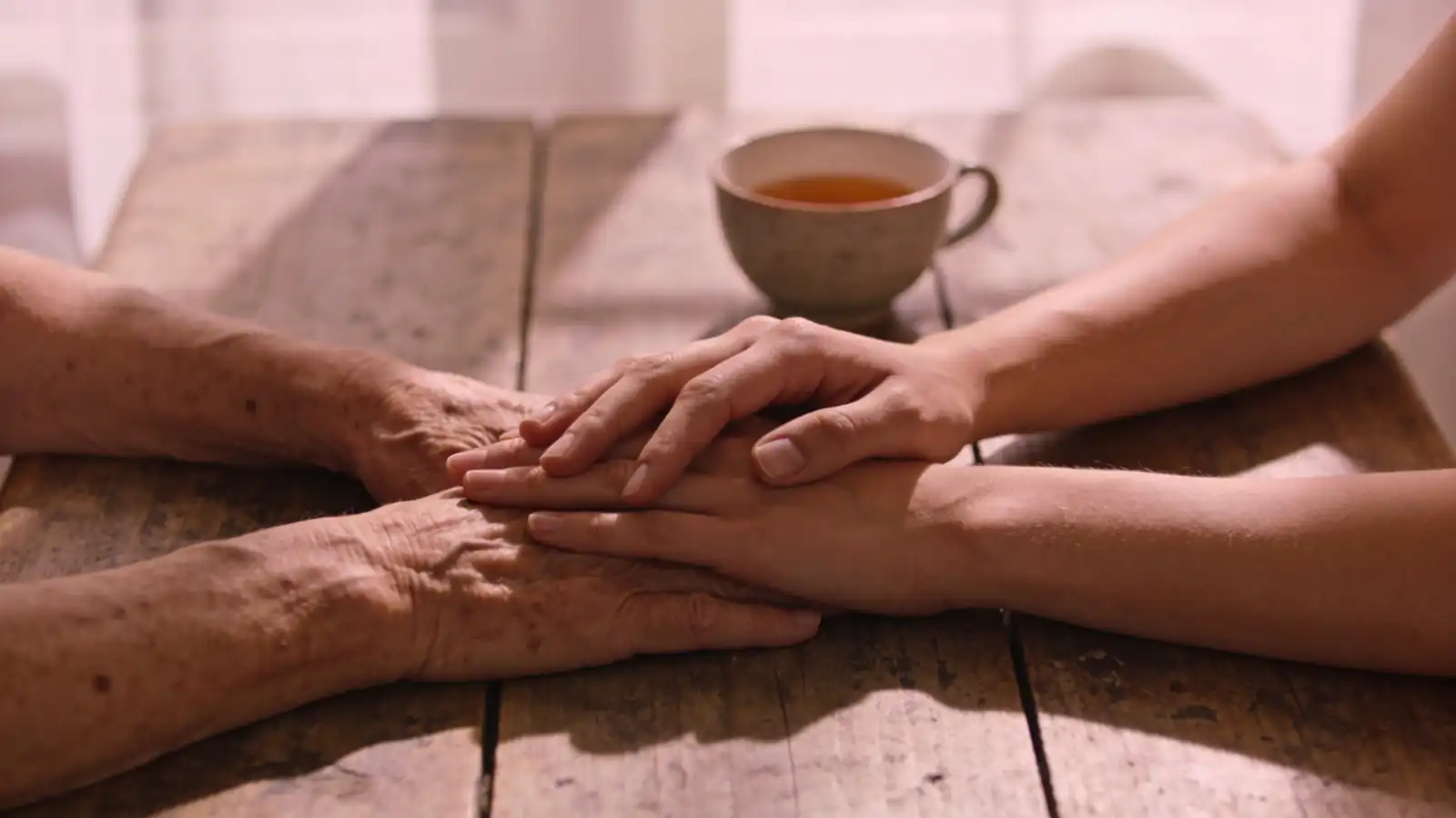 Elderly hands held gently in younger hands across a wooden table — the quiet love between generations at a time of grief